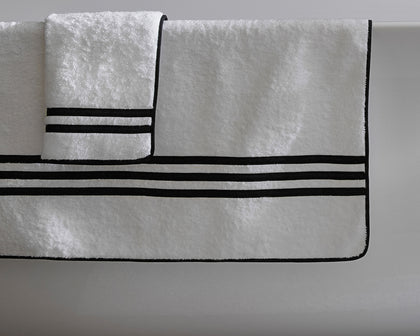 White Newport towels with black striped borders hanging from a tub edge.