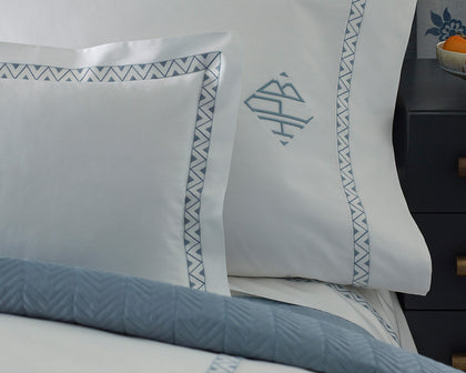 White Prado bedding with blue zigzag embroidered trim and monogram, styled on a bed with a dark nightstand.