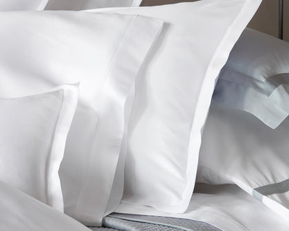 White Talita Satin Stitch pillow shams layered with blue-trimmed pillows on a bed.