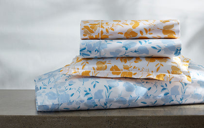 Alexandra floral print sheets in gold and blue, stacked on a stone ledge.