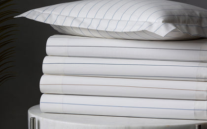 White Amalfi pinstripe sheets stacked on a marble table beside a palm frond.