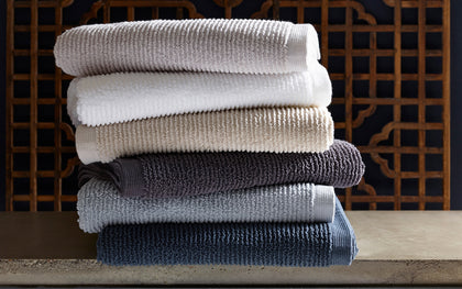 Ribbed Aman towels stacked in white, tan, gray, and navy, in front of a carved wood screen.