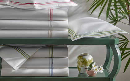 White Amara Triple Pearl sheets with triple-dot embroidered trim in assorted colors, stacked on a green table.