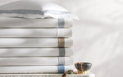 White Ambrose sheets with satin bordered trim in blue and tan, stacked on an inlaid dresser.