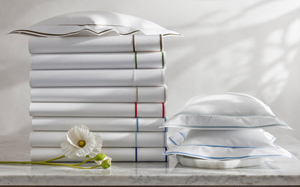 White Ansonia sheets with single-stripe trim in assorted colors, stacked beside a white poppy.