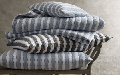 Apollo Stripe Matelasse quilted bedding in blue and gray, stacked on a wooden bench.