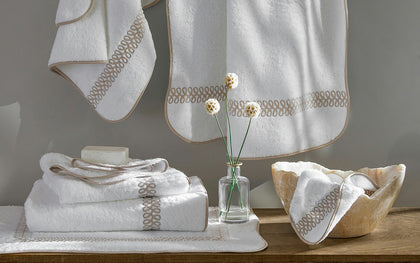White Astor Braid towels with tan loop-embroidered trim, hung and stacked with a bud vase.