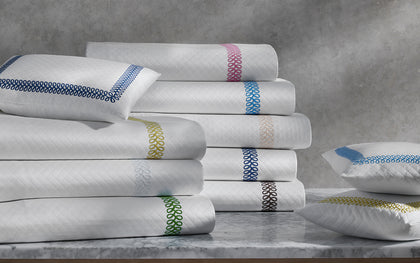 White Astor Braid quilted bedding with loop-embroidered trim in a rainbow of colors, stacked on a marble table.