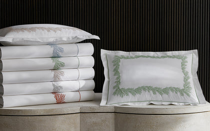 White Atoll sheets with coral-branch embroidered trim in assorted colors, stacked beside a green-embroidered sham.