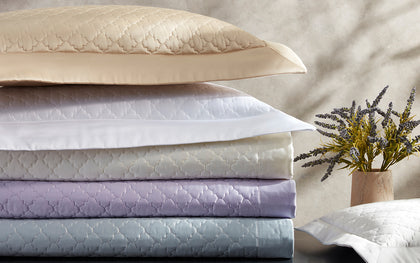 Ava quatrefoil-quilted bedding stacked in gold, white, lavender, and blue, beside a sprig of lavender.