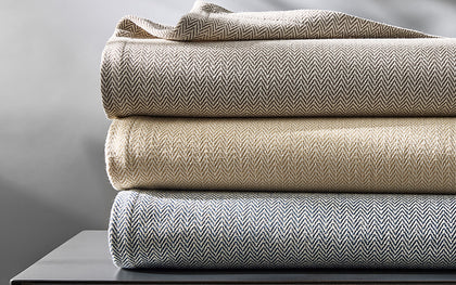 Avalon herringbone-woven blankets folded in gray, tan, and blue, stacked on a dark table.