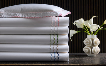 White Axel sheets with wavy embroidered trim in assorted colors, stacked beside calla lilies.