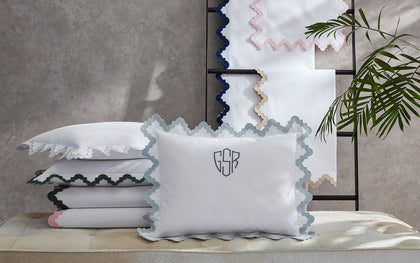 White Aziza pillow with blue wavy trim and a monogram, stacked with sheets in assorted colors.