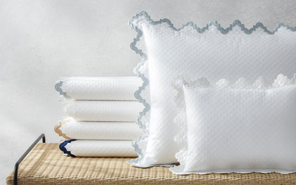 White Aziza Matelasse quilted bedding with gray wavy trim, stacked on a woven bench.