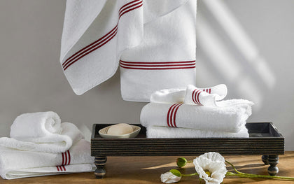 White Bel Tempo towels with red triple-stripe trim, hung and stacked beside a poppy flower.