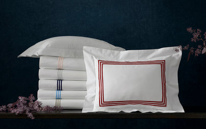 Stacked white Bel Tempo sheets with colorful striped trim beside a red-bordered euro sham.