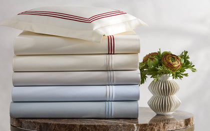 Stacked Bel Tempo Nocturne sheets with striped embroidered trim in ivory and pale blue, beside a vase of flowers.