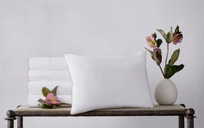 Stacked white Bryant sheets with colored piped trim, beside a tan-piped pillow and a pink flower.