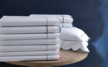 White Butterfield scalloped sheets stacked with colored trim, beside a navy-trimmed sham.