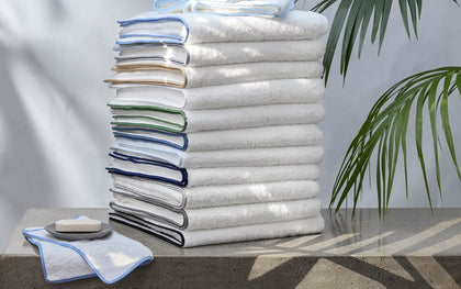 Tall stack of white Cairo towels with piped trim in a rainbow of colors, beside soap and a palm plant.