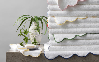 White Cairo Scallop towels with wavy scalloped trim in assorted colors, stacked beside soap and a plant.