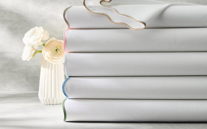 White Camilla Pique sheets stacked with colored edge trim, beside a vase of white ranunculus.