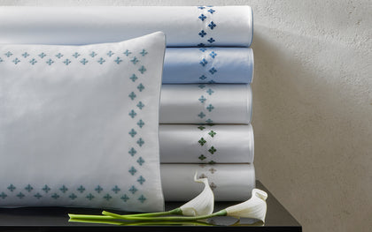 White Casablanca sheets with diamond cross-stitch trim in assorted colors, stacked beside calla lilies.