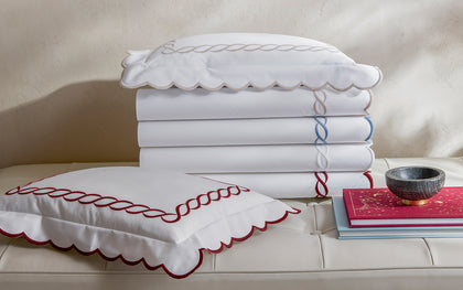 White Classic Chain Scallop sheets with scalloped chain-embroidered trim in tan, blue, and red, stacked on a tufted bench.
