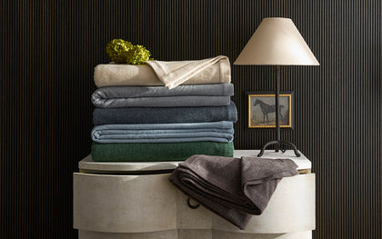 Cosmo blankets folded in tan, gray, navy, and green, stacked on a chest beside a horse painting.