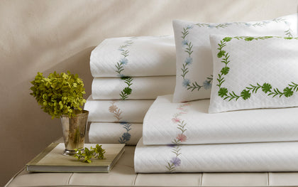 White Daphne Matelasse quilted sheets with floral embroidery, stacked on a bench beside a plant.
