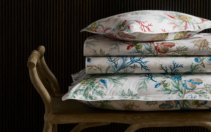Del Tesoro coral and shell print sheets in red and blue, stacked on a wooden bench.