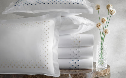 White Dorothy sheets with polka-dot embroidered borders, stacked beside a vase of dried flowers.