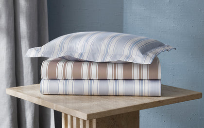 Striped Elio sheets in tan and blue, stacked on a stone side table.