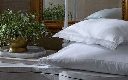 White Grace pillows with lace trimmed edges on a bed near a mirror and flowers.