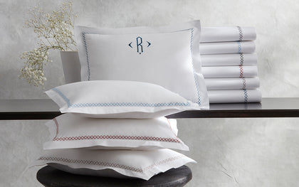 White Hatch pillows and rolled sheets with cross-stitch trim in assorted colors, beside a monogrammed sham and baby's breath.