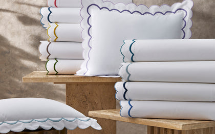 White India scalloped sheets and shams stacked on stone tables in a rainbow of trim colors.
