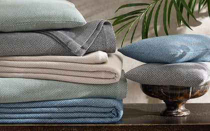 Stacked Jackson houndstooth-textured sheets and pillows in sage, black, tan, and blue, beside a palm frond.