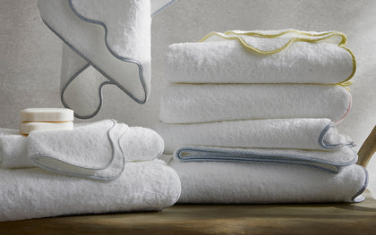 White Jasper towels with braided trim in yellow, gray, and pink, stacked on a wood bench.