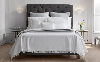 White Lace Pique bedding with floral lace trim, styled on a bed with a gray tufted headboard.
