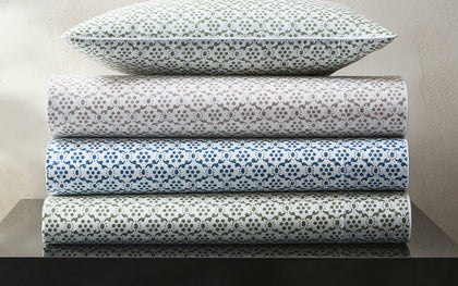 Laksha medallion print sheets stacked in green, tan, blue, and olive, topped with a calla lily.