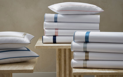 White Louise sheets and pillows stacked with satin bordered trim in assorted colors.