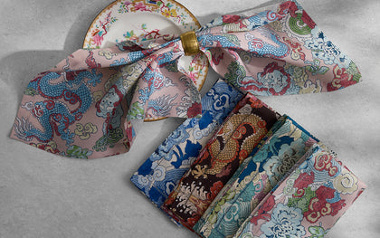 Magic Mountain dragon print napkins in pink, blue, and gold, folded with a napkin ring beside a floral plate.