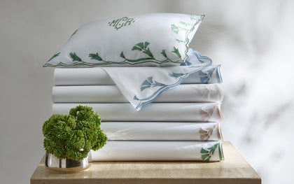 White Marcella sheets with vine-embroidered scalloped trim in assorted colors, stacked with a monogrammed pillow.