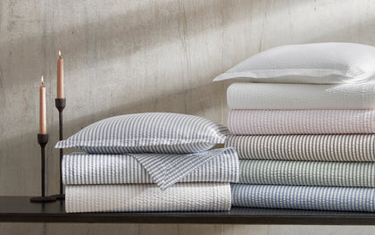 Blue-striped Matteo bedding folded on a dark shelf beside lit taper candles, with striped bedding in assorted colors stacked alongside.