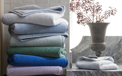 Milagro bath towels stacked in assorted colors beside a folded towel on marble with a dried berry arrangement.