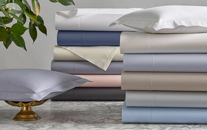 Milano Hemstitch sheets stacked in assorted colors on marble beside a lavender pillow in a brass stand.