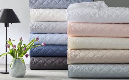Milano Quilt bedding stacked in assorted colors beside a marble vase of tulips and a black lamp.
