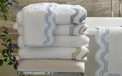 White Mirasol towels with scalloped blue, gray, and gold trim stacked beside purple flowers.