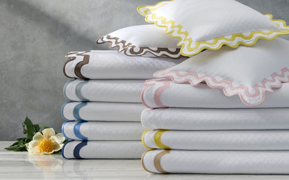 White Mirasol Matelasse coverlets with scalloped multicolor trim stacked with shams beside a white flower.