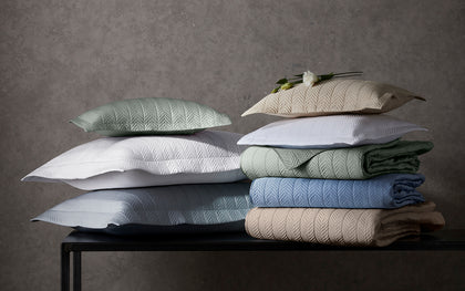 Netto herringbone-quilted shams and bedding stacked in assorted colors on a black console.